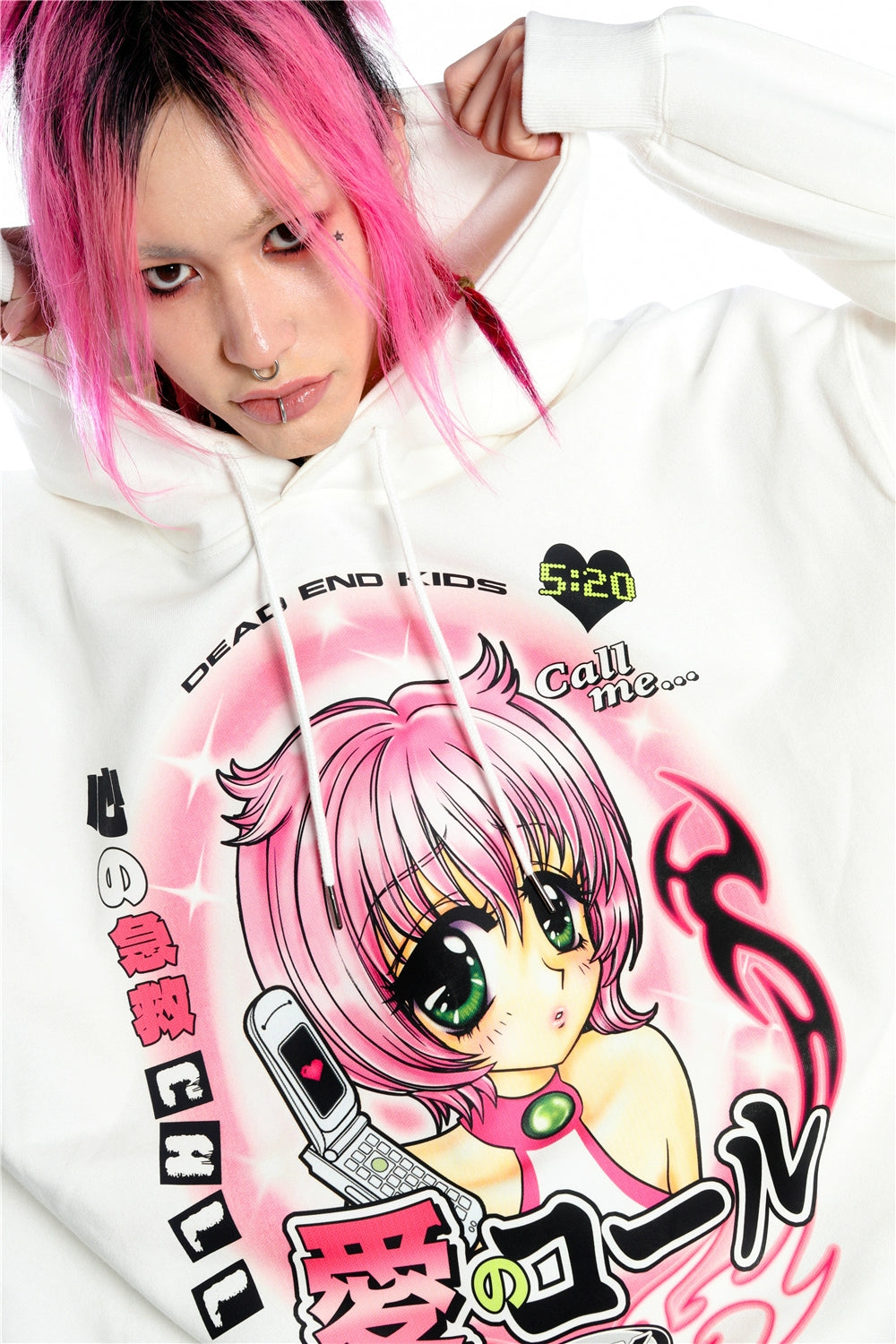 A person with pink and black hair wears the Seakoff Y2K Aesthetic Anime Hoodie, featuring a kawaii-style manga girl graphic. The white pullover shows a pink-haired character holding a phone amidst Y2K text elements, as the person looks directly at the camera.