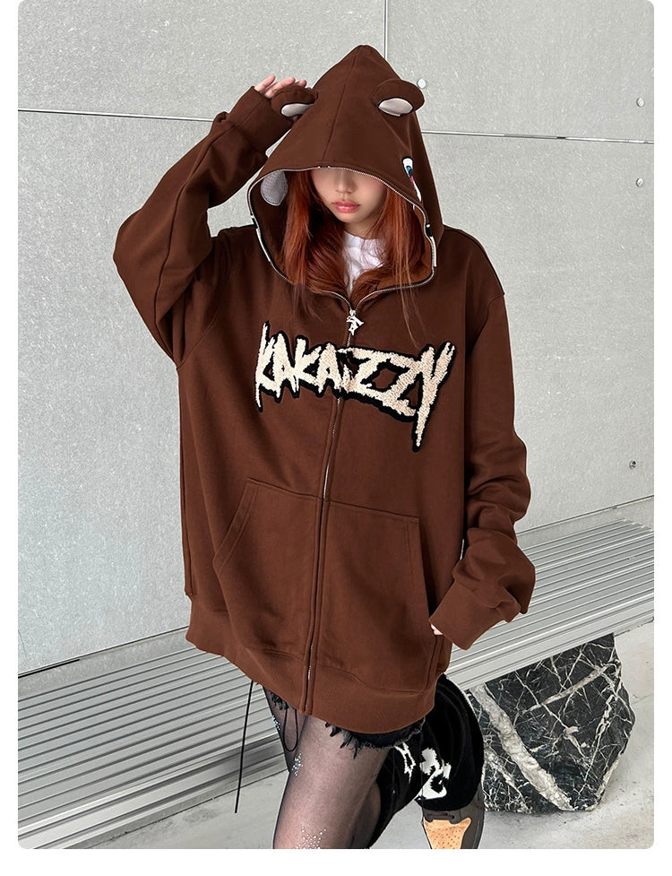 A person in a Seakoff Cute Bear Hoodie (400GSM oversized brown zip-up with ears) stands against a gray wall, paired with black lace-patterned tights and shoes, exuding a cool streetwear vibe.