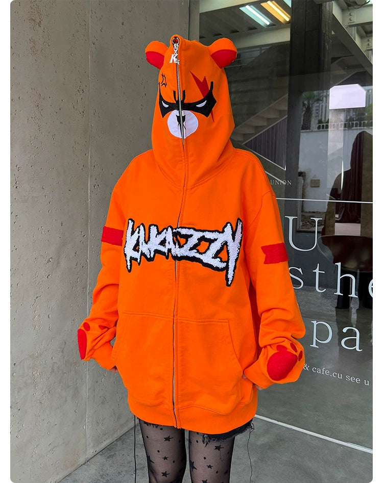 A person wears the Seakoff Furious Orange Bear Hoodie in premium 400GSM cotton, featuring an angry bear face with ears and "KRAZZY" text. Paired with black star-patterned tights and fingerless gloves, they stand against a concrete wall with partial signage in the background.