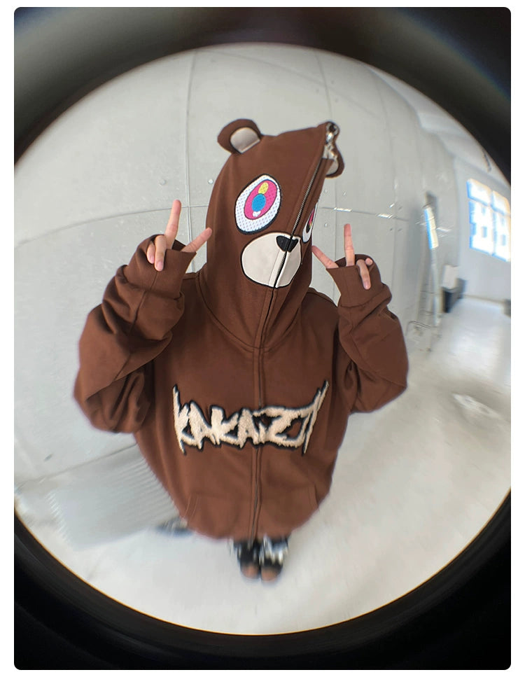 A person wearing the Seakoff Cute Bear Hoodie, an oversized 400GSM brown zip-up streetwear sweatshirt with bear ears, gives peace signs. The "KAKAZY" lettering adds flair, all captured in a playful fisheye lens effect.