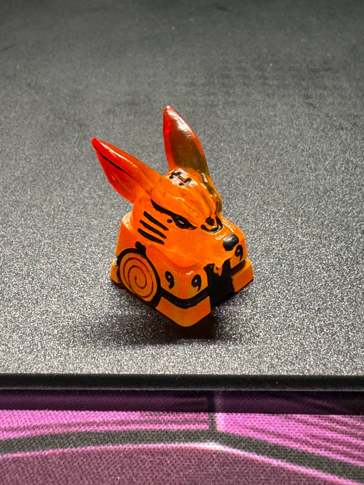 A small, orange figurine with red-tipped ears and black markings, resembling a stylized fox, sits on a dark surface. The figure has distinct detail lines and patterns.