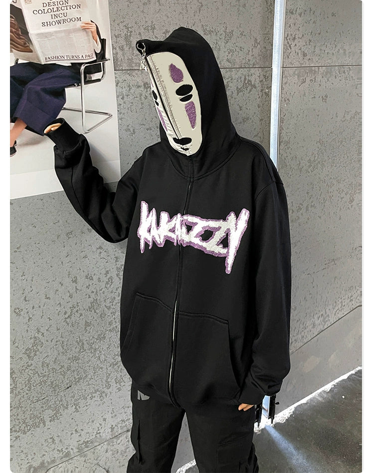 Against a gray textured wall, someone in the Spirited Away "No-Face Anime Hoodie," a 400GSM oversized zip-up sweatshirt with white and purple "MAKIZZY" text, wears a white mask featuring purple markings and a vertical zipper, capturing the mysterious vibes reminiscent of No-Face.
