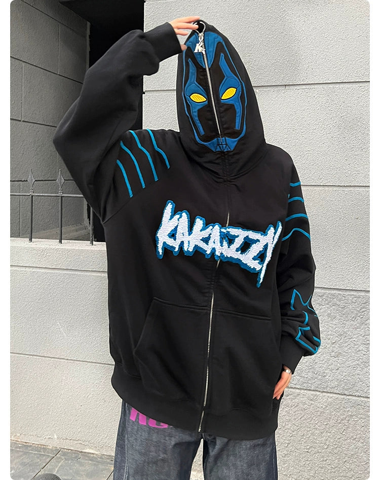 A person in a Seakoff Cyberpunk Superhero-Inspired Hoodie stands against a gray wall. The oversized black 400GSM zip-up features "Kakazzzi" on the front and stylized blue and yellow mask-like details on the hood, embodying futuristic allure perfect for sci-fi fans.