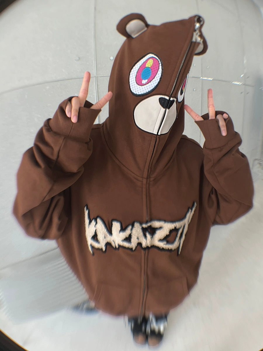 A person is rocking a Seakoff Cute Bear Hoodie, an oversized 400GSM brown zip-up sweatshirt with ears. The hoodie is zipped over their face, displaying "KAKAZE" in stylized letters as they flash peace signs, perfectly capturing the streetwear vibe.