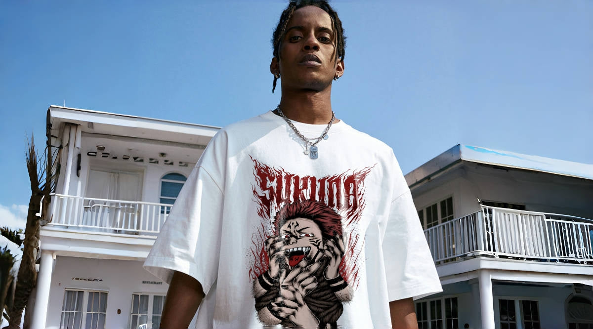 A person stands in front of a two-story white building with balconies. They are wearing a white graphic T-shirt featuring a detailed illustration and are looking directly at the camera. The sky is clear and blue.