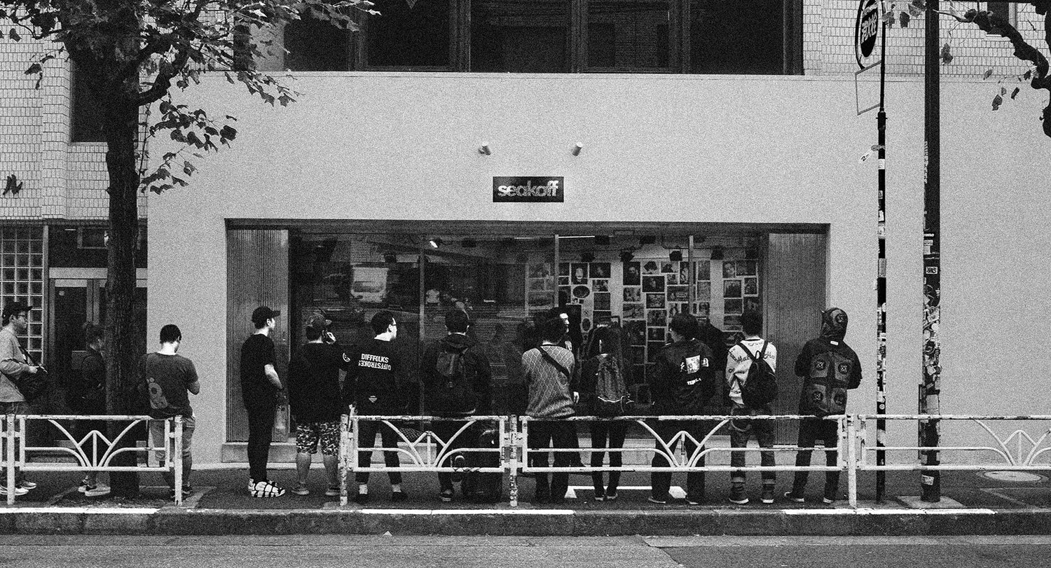 A group of people stand outside a shop with a glass front displaying various items and photos. The building is in a city environment with trees and a metal fence along the sidewalk. The image is in black and white.