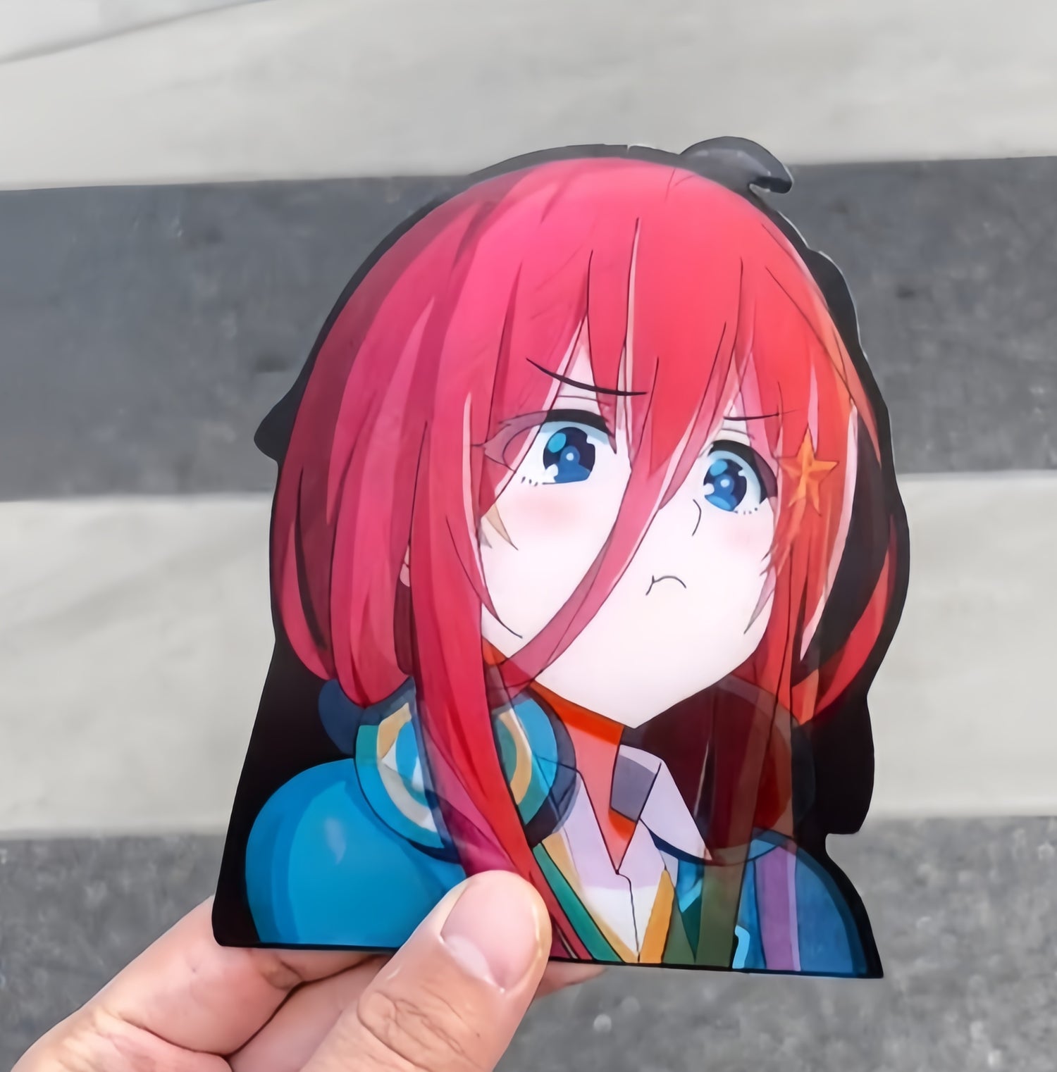 A sticker of an animated character with long red hair, blue eyes, and a small star hair accessory. The character has a pouting expression and wears a blue outfit. A hand holds the sticker against a blurred background.