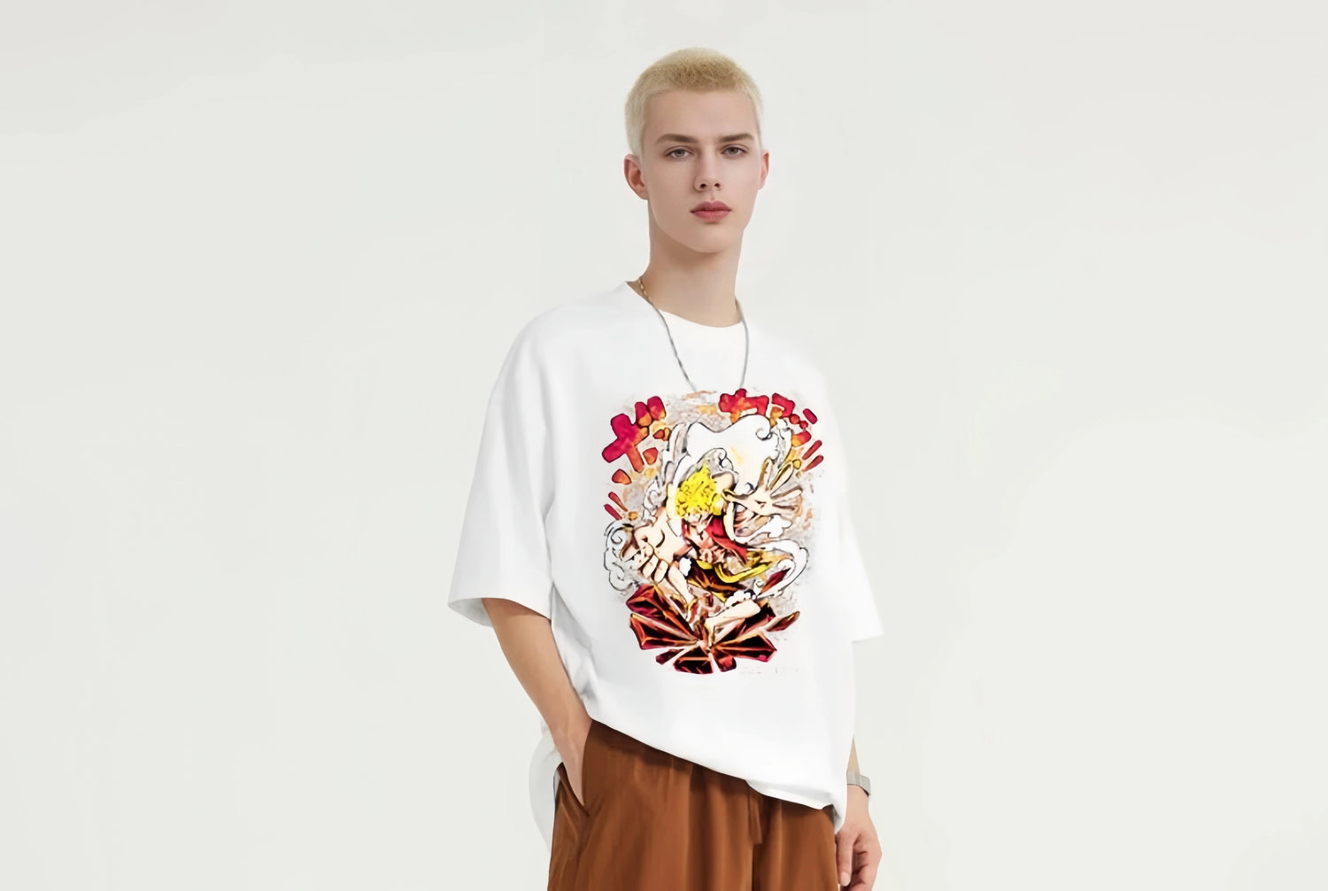 A person with short blond hair wearing a white t-shirt featuring a colorful graphic and brown pants stands against a plain white background. They have one hand in a pocket and are looking ahead.