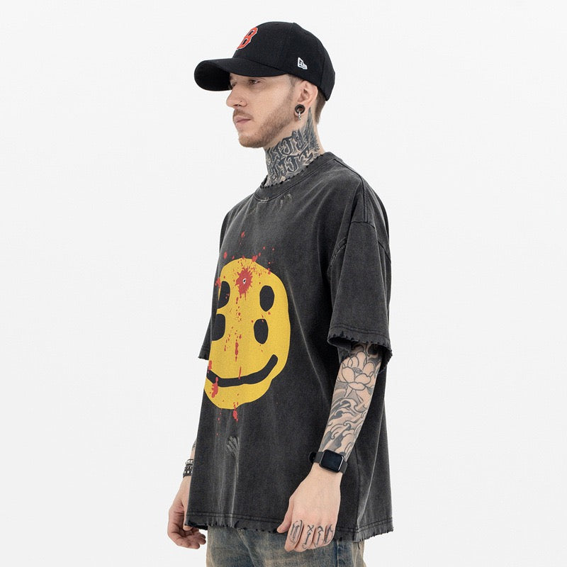 Oversized washed black graphic tee with distressed yellow smiley face print on a model with tattoos and cap