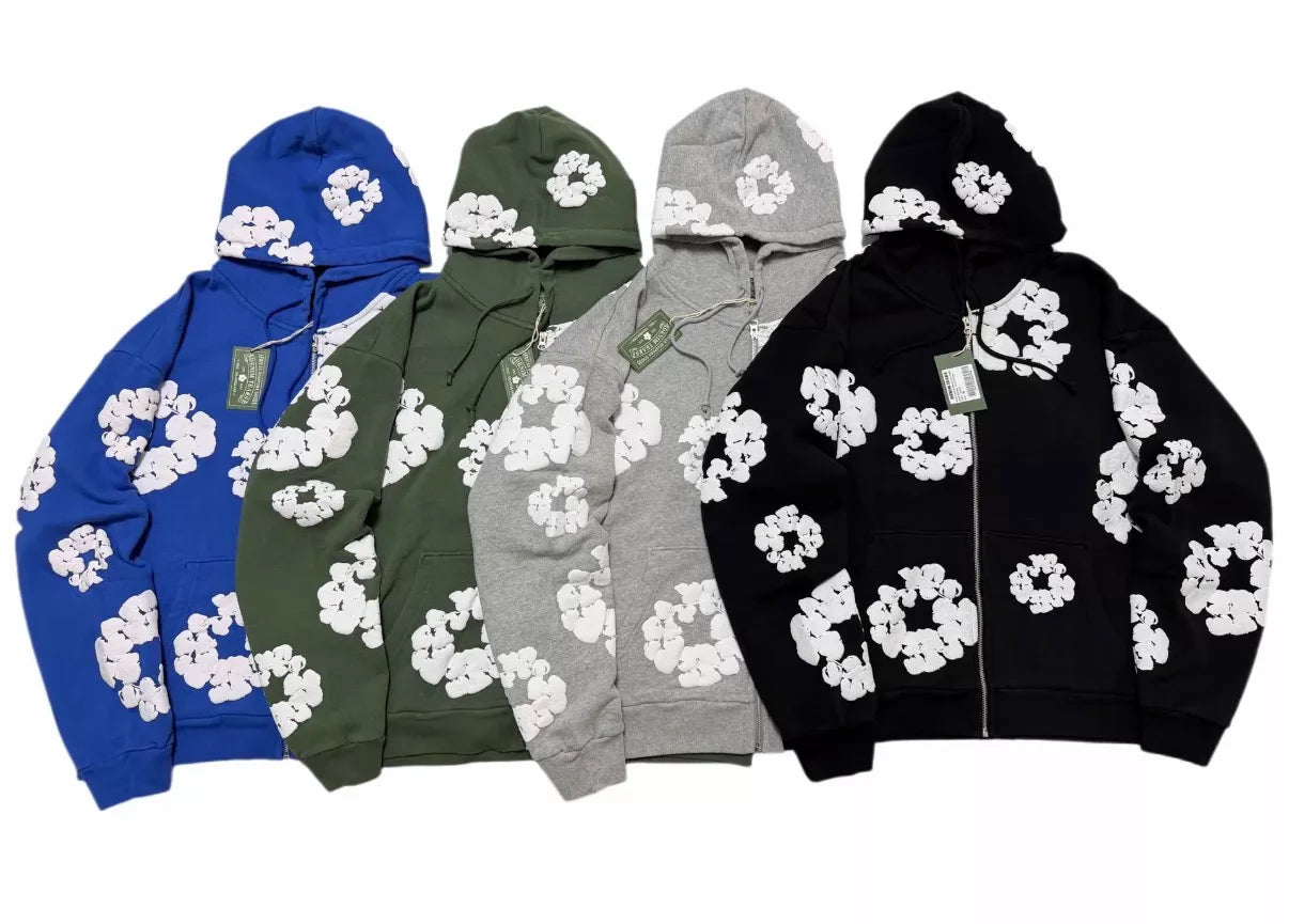Four Denim Tears hoodies in blue, green, gray, and black colors, each covered with white flower designs and featuring front zippers and hanging tags.