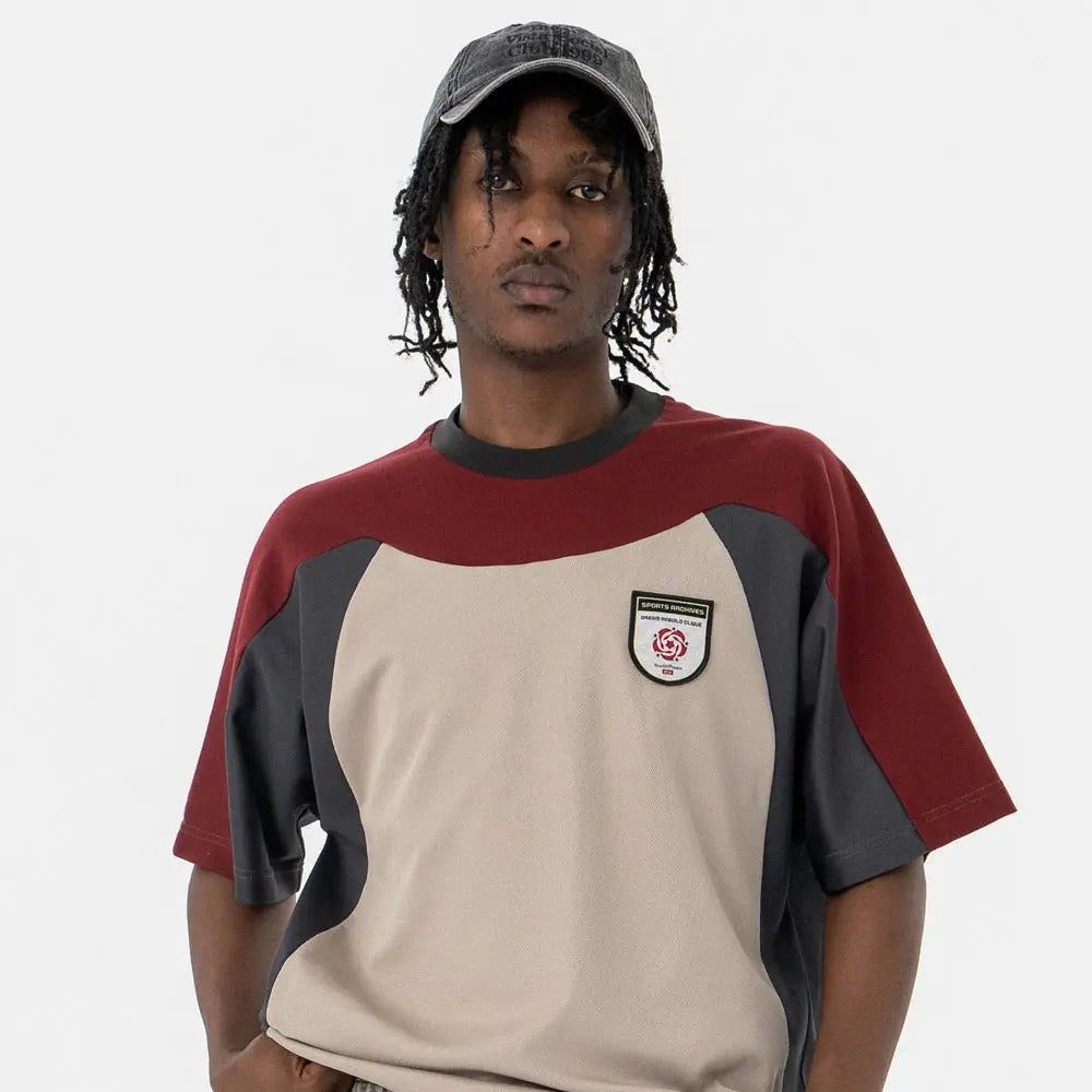A person wearing a gray cap and a loose beige, maroon, and black color-blocked shirt with a patch on the chest stands against a plain white background.