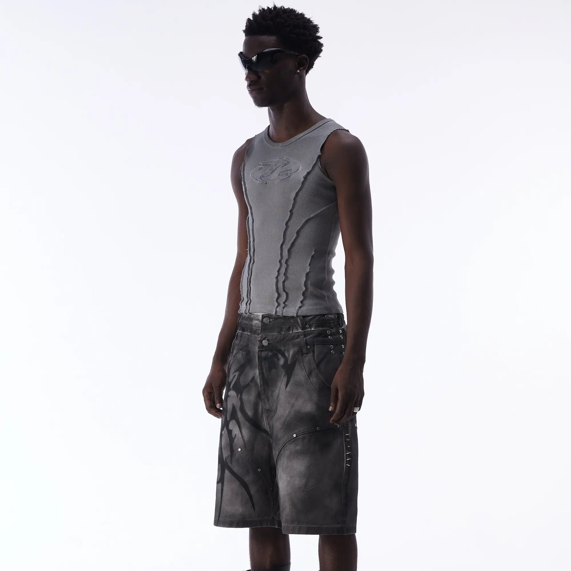 A man wearing sunglasses, a fitted gray tank top, and loose, knee-length black and gray shorts stands against a plain white background.