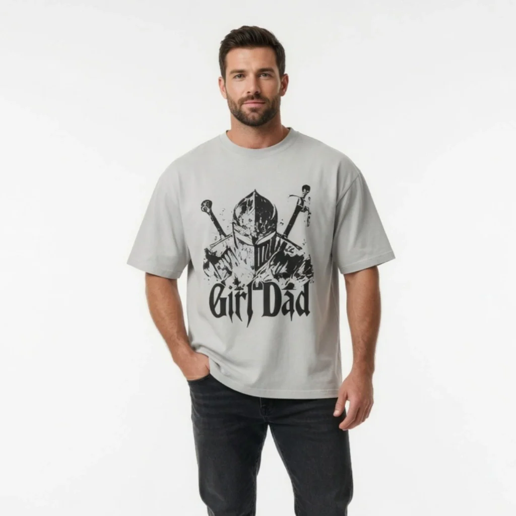 A man with short dark hair and a beard wears a light gray T-shirt featuring a medieval knight graphic with two swords and the words Girl Dad. He stands facing forward with one hand in his pocket against a plain white background.