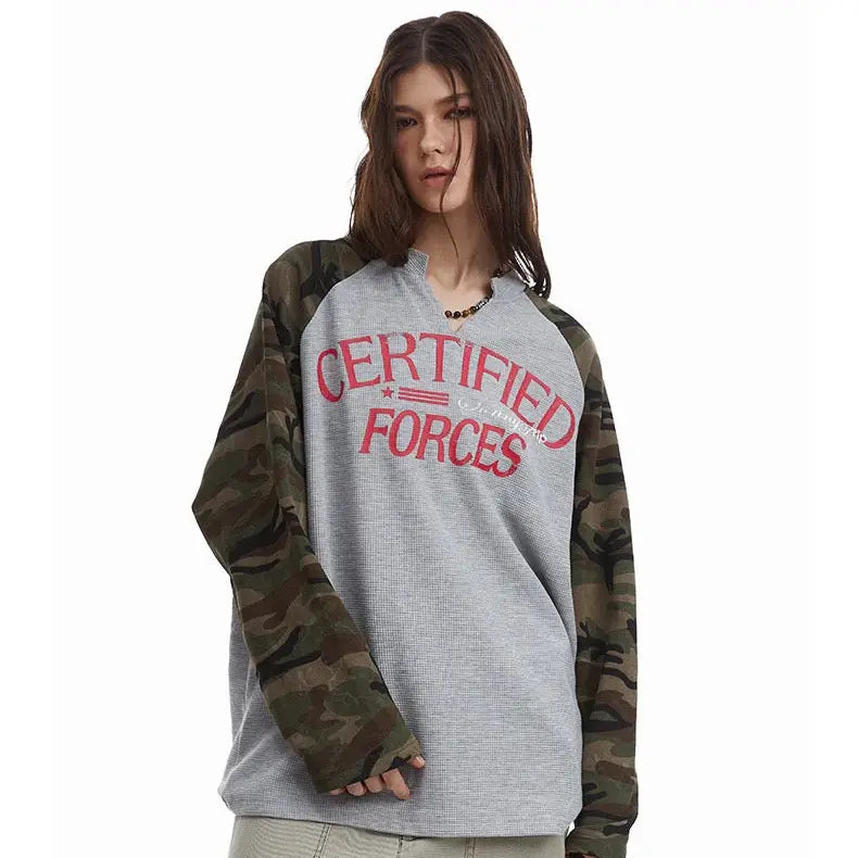 A young woman with long brown hair wears a gray shirt with Certified Forces in red text and camouflage-patterned sleeves, standing against a plain white background.
