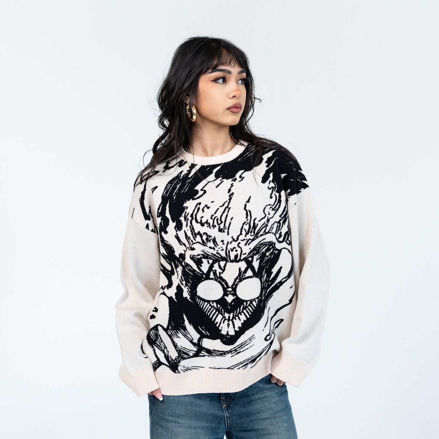 A person with long dark hair stands in a light sweater featuring a bold black and white graphic design on the front. They are wearing jeans and looking to the side, set against a plain white background.