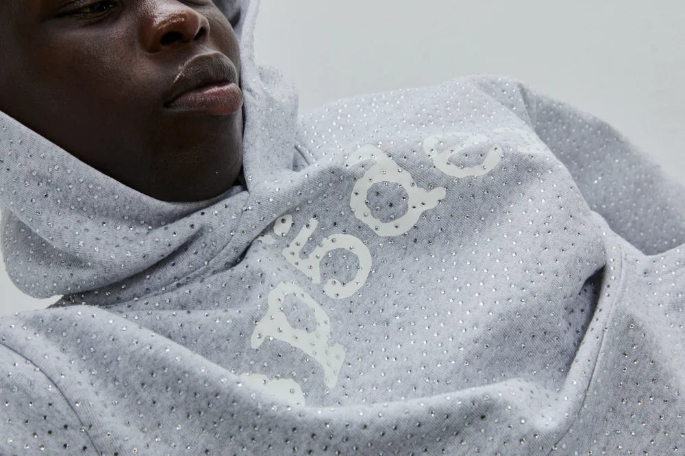 A person wearing a light gray sp5der hoodie with sparkling embellishments and white text partially visible on the front, lying back and looking downward.