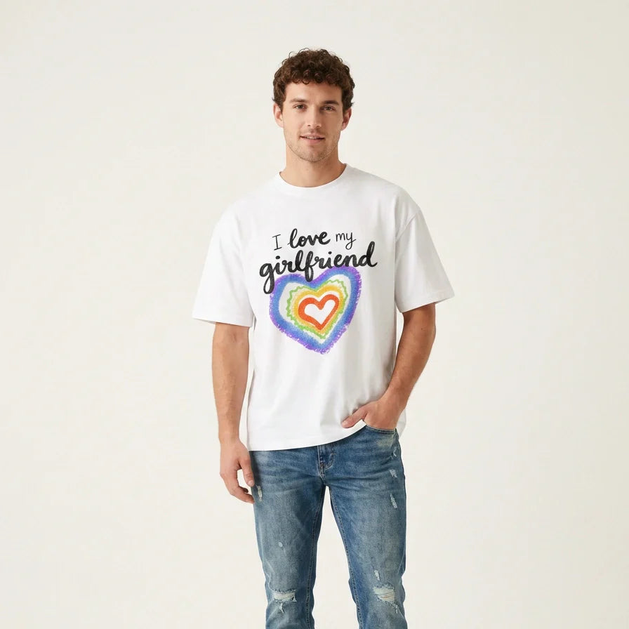 A man stands against a plain background, wearing a white T-shirt that says I love my girlfriend above a colorful, rainbow heart design. He is also wearing blue jeans and has one hand in his pocket.