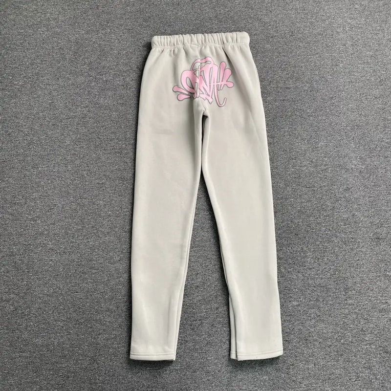 Syna World Cream Joggers with Pink Drawstring, exuding streetwear vibes, are showcased laid flat on a gray textured surface.