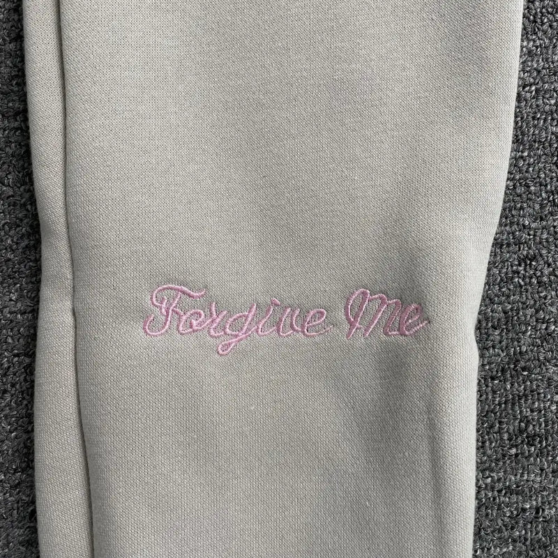 Close-up of light gray fabric with "Forgive Me" in pink embroidery on a dark gray background—a subtle streetwear-inspired detail ideal for pairing with the Syna World Cream Joggers with Pink Drawstring.