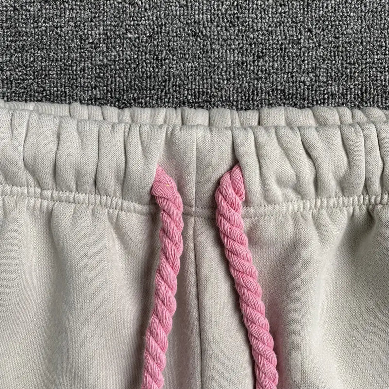 Close-up of Syna World Cream Joggers with Pink Drawstring, shown on a gray carpet—featuring an elastic waistband and bold pink drawstrings for a standout streetwear style.