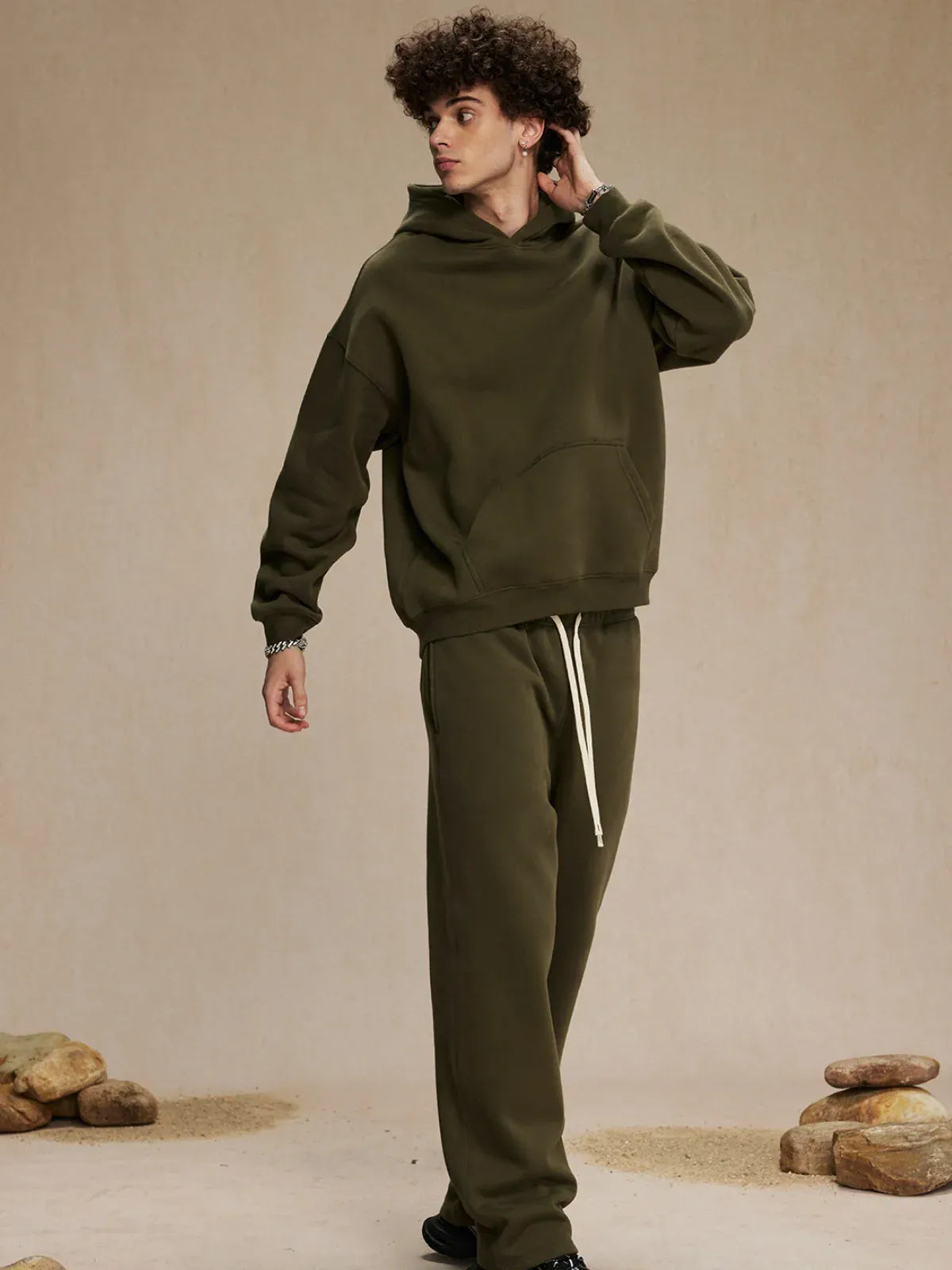 Someone wearing the Custom Anime Character Olive Green Hoodie—Personalized 350GSM 100% Cotton Pullover—and matching sweatpants stands indoors on a beige floor with scattered rocks.