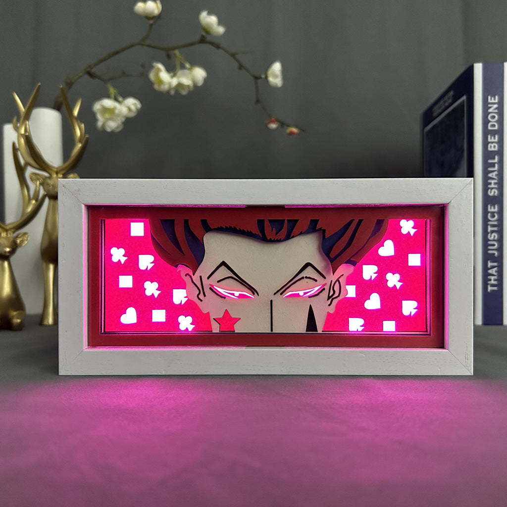 The Hunter × Hunter Anime Light Box features Hisoka with a 3D effect. It displays his eyes and forehead amid card suit symbols on a bright pink backdrop, complemented by decorative items like deer figurines and a book. The light box includes a 16-color adjustable LED light for added flair.