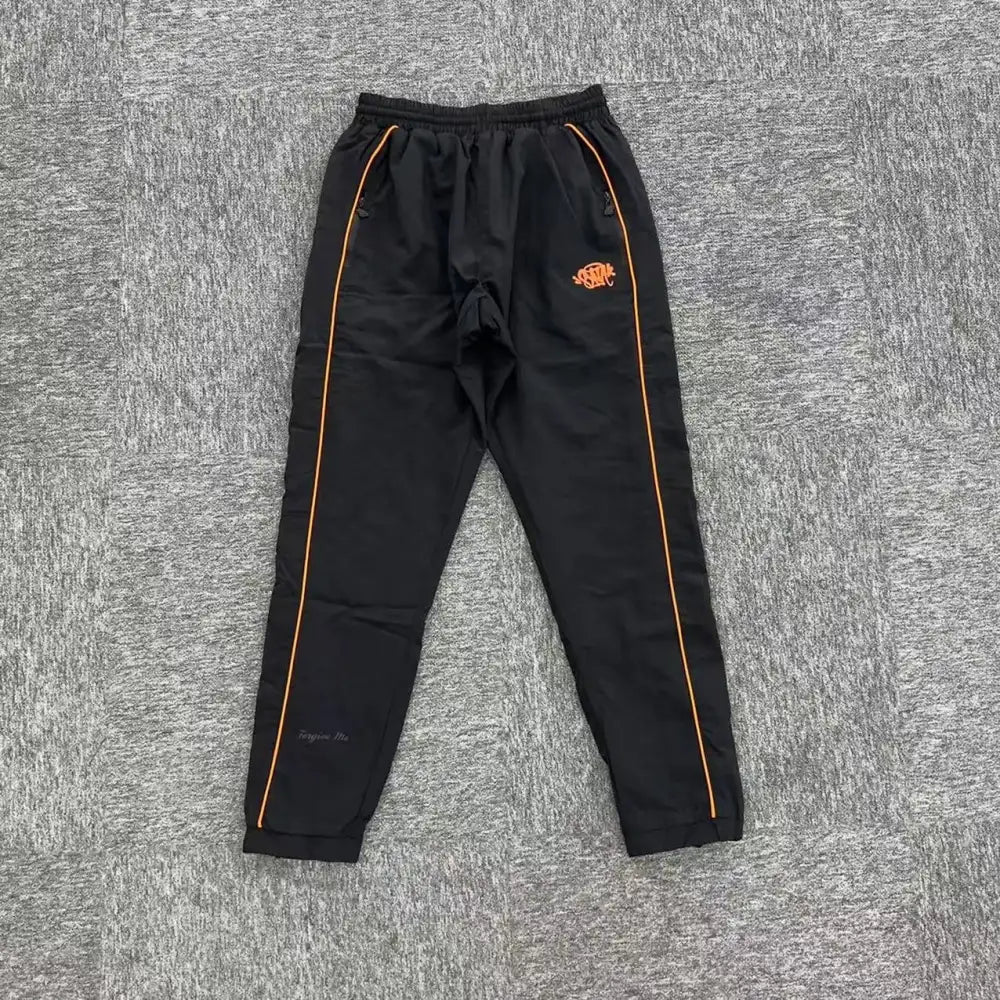 Black jogger pants with orange side piping and embroidered graphic on left hip, laid flat on grey surface