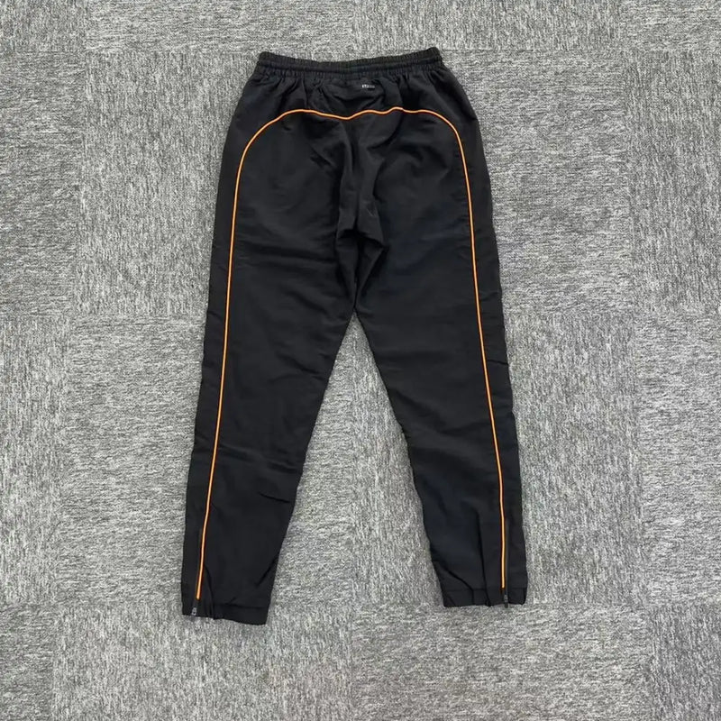 Syna World Black Orange Piping Jogger Pants, crafted from a cotton-polyester blend with orange side piping, are displayed flat on a gray textured surface.