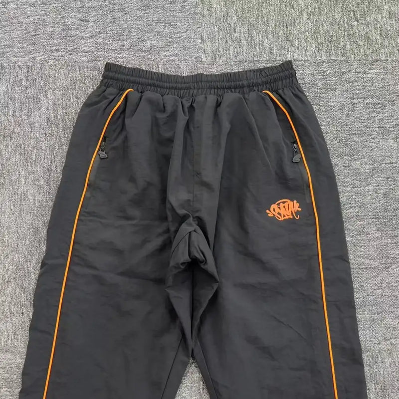 The Syna World Black Orange Piping Jogger Pants feature a comfy cotton-poly blend, orange side piping, elastic waistband, zippered side pockets, and an orange graphic logo on the left thigh. Shown laid flat on a gray carpet.