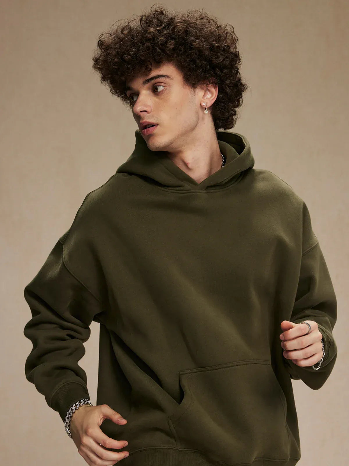 A young man with curly hair models the Custom Anime Character Olive Green Hoodie—Personalized 350GSM 100% Cotton Pullover—with one hand slightly raised, looking to his left against a plain beige background.