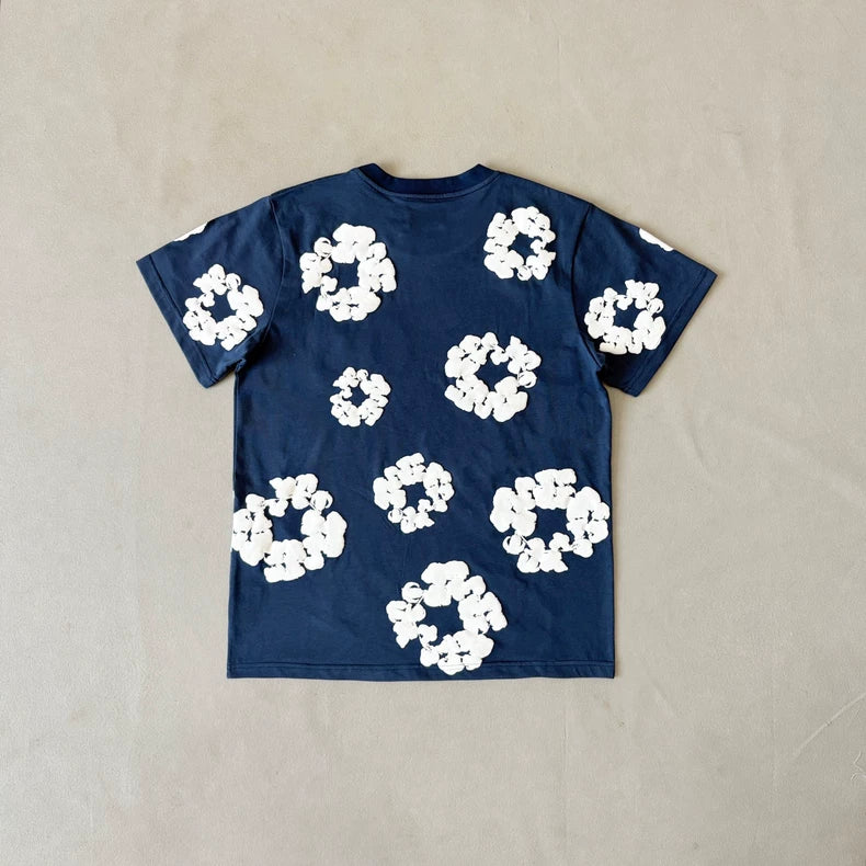 The Denim Tears Navy Cotton Wreath All-Over Print T-Shirt, shown flat on a beige surface, features an oversized fit and is decorated with irregular white flower-like patterns across the back.
