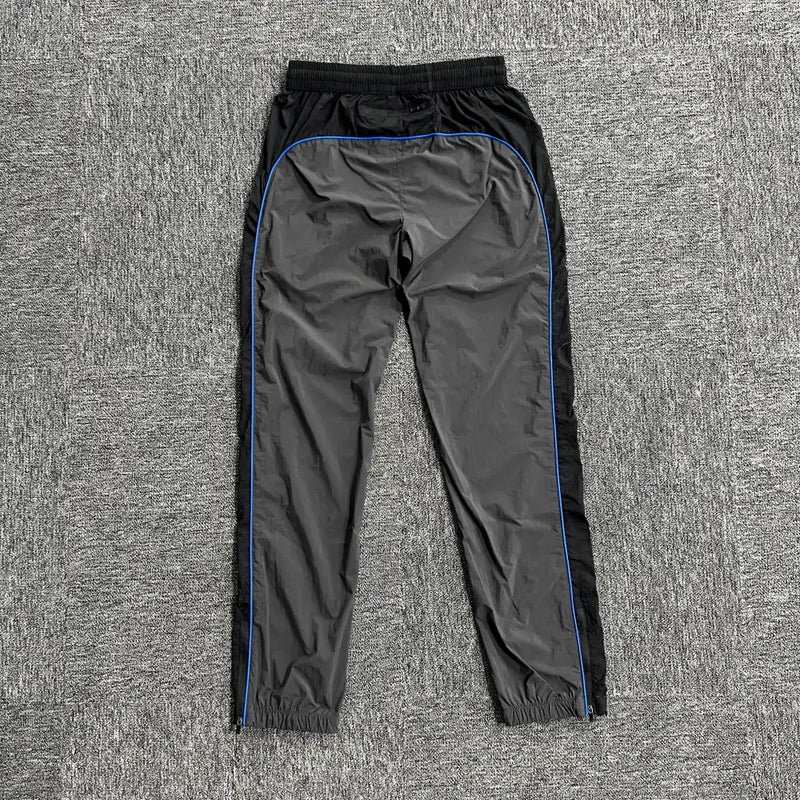 The Syna World Grey Black Patchwork Track Pants, featuring black side panels and blue piping, are displayed flat on a textured gray surface.