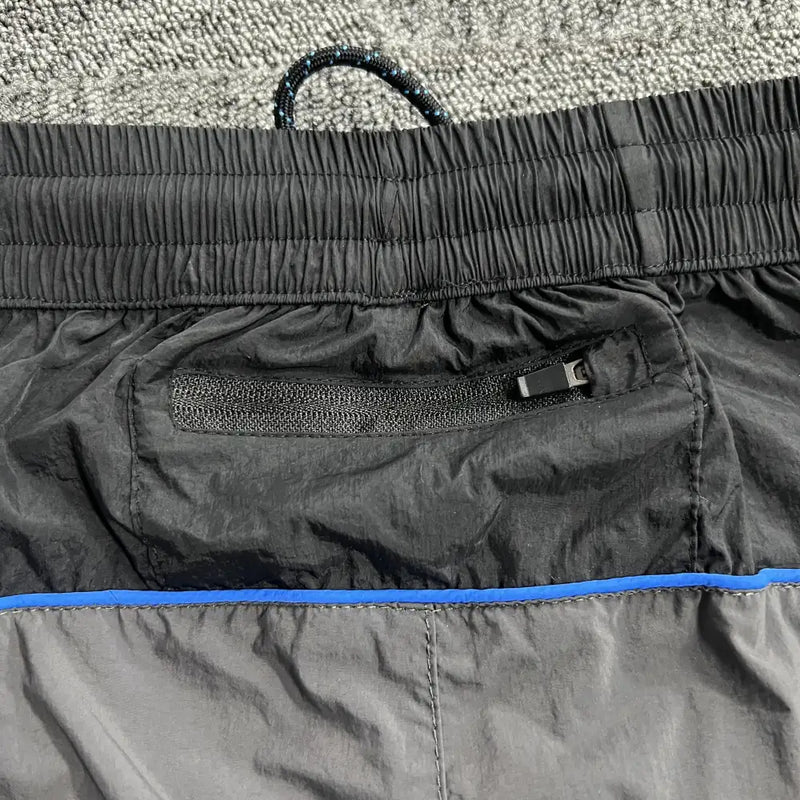 Close-up of Syna World Grey Black Patchwork Track Pants with an elastic waistband, black zipper pocket, blue trim detail, and drawstring, displayed on a textured grey carpet.