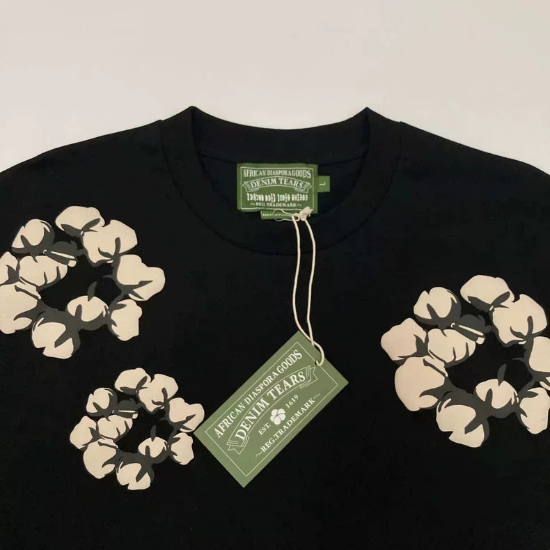 The Denim Tears Black Cotton Wreath Allover Print T-Shirt features white floral and cotton wreath graphics on the chest and shoulders, plus a green tag with "AFRICAN DIASPORA GOODS SEWING TEARS" in white text.