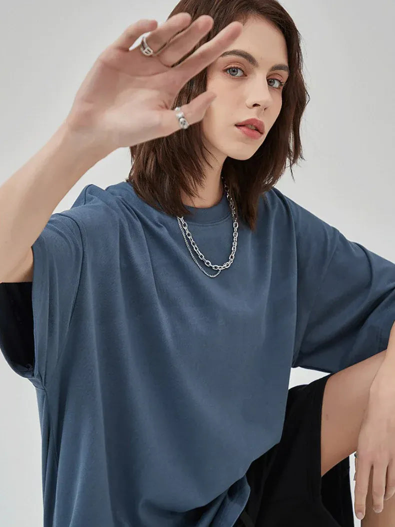 Individual with shoulder-length brown hair wears a Custom Anime Character Steel Blue T-Shirt - Personalized 230GSM 100% Cotton Tee, paired with a silver chain necklace and rings, seated and raising one hand towards the camera.