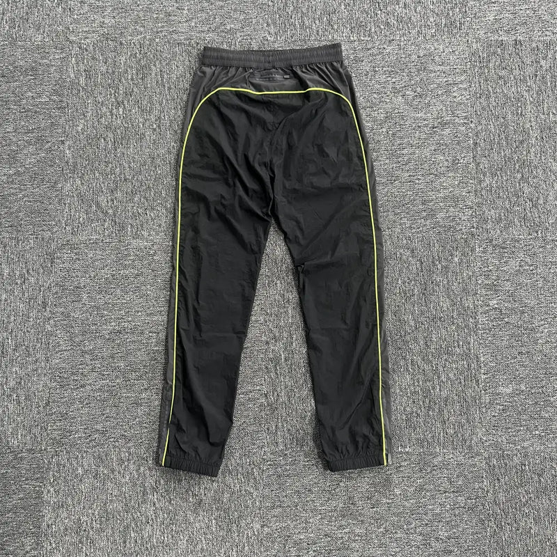 Syna World Black Yellow Logo Track Pants, featuring yellow piping, an elastic waistband, and cuffed ankles for a comfy, stylish fit, shown laid flat on a gray textured carpet.