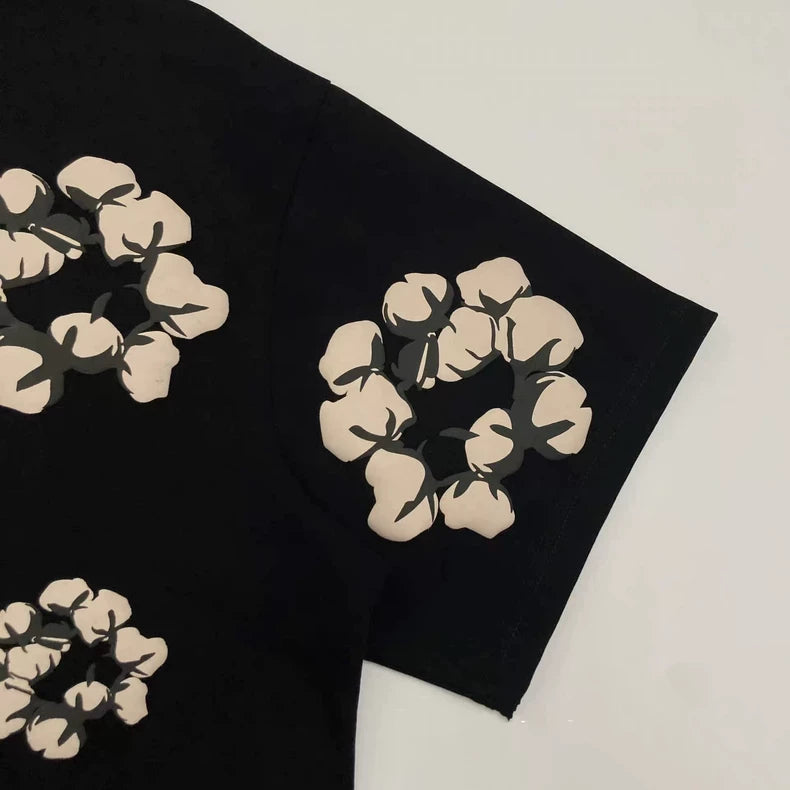 The Denim Tears Black Cotton Wreath Allover Print T-Shirt features beige, flower-like cotton wreath graphics in circular patterns on the sleeves and body, set against a black background.