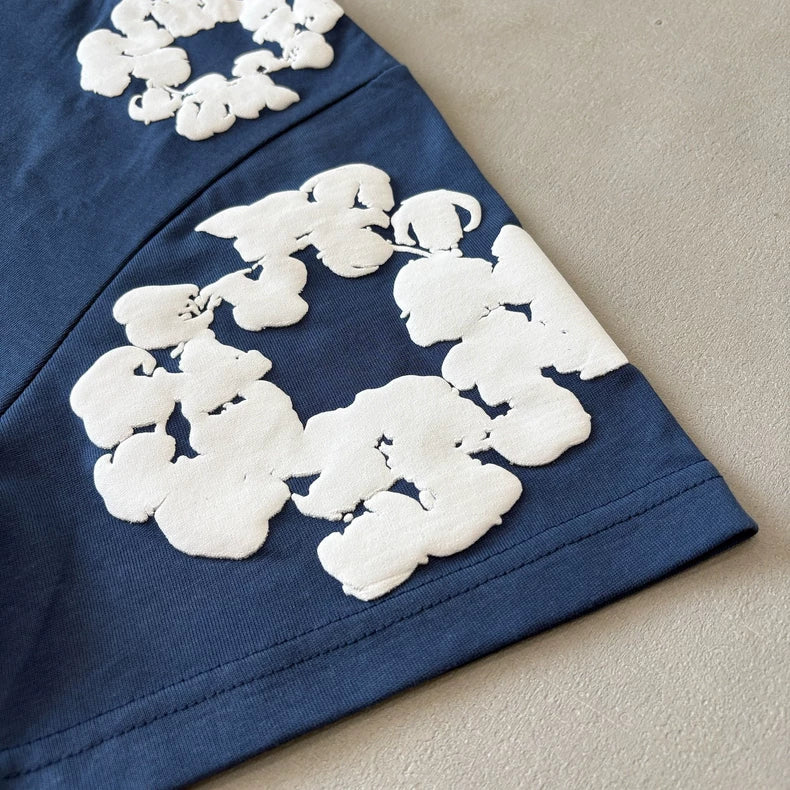 Close-up of blue fabric with white floral appliqué, echoing the cotton wreath graphic featured on the Denim Tears Navy Cotton Wreath All-Over Print T-Shirt, shown on a neutral surface.