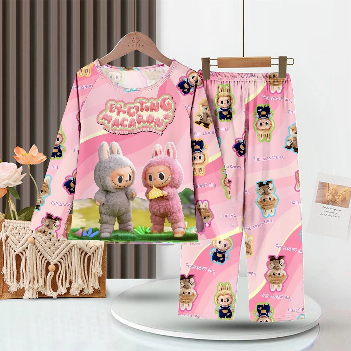 Displayed on a hanger, the LABUBU Baby Pajama Set from the &quot;Exciting Macaron&quot; series features pink milk silk fabric with cartoon animals and &quot;Exciting Vacation&quot; text. The pants have smaller patterns, set against a cozy rug and flowers backdrop.