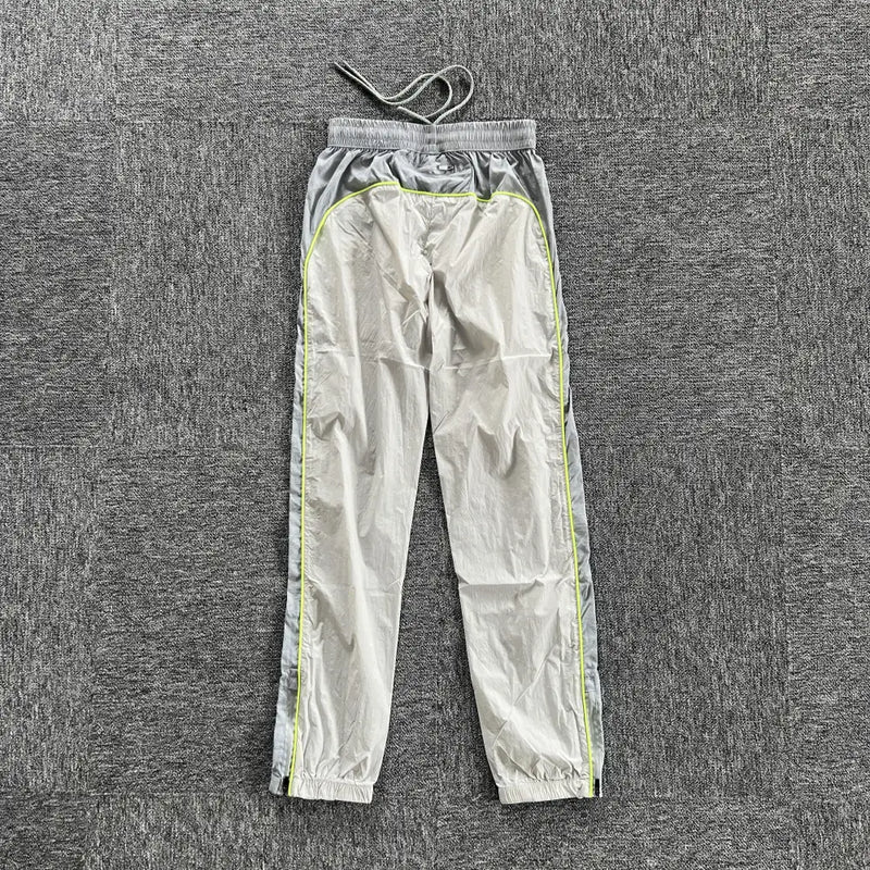 Syna World Grey Yellow Logo Track Pants featuring elastic cuffs, a drawstring waistband, zip pockets, and neon yellow piping are laid flat on a textured gray surface.