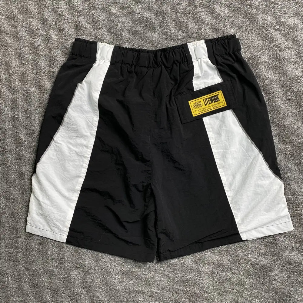 Corteiz Black Colorblock Nylon Shorts with Belt, made of nylon, feature vertical white side stripes, an elastic waistband, and a yellow text patch on the right. Shown laid flat on a gray textured surface.