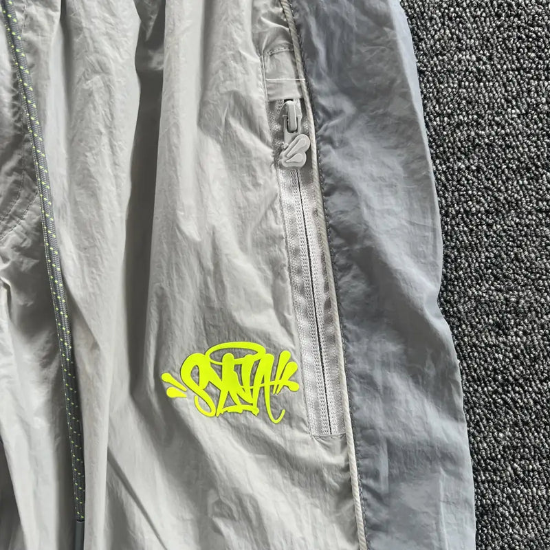 A close-up of Syna World Grey Yellow Logo Track Pants with zip pockets and neon yellow graffiti-style "SNA" text near the pocket, laid on a dark gray textured surface—ideal for pairing with athleisure or matching tops.
