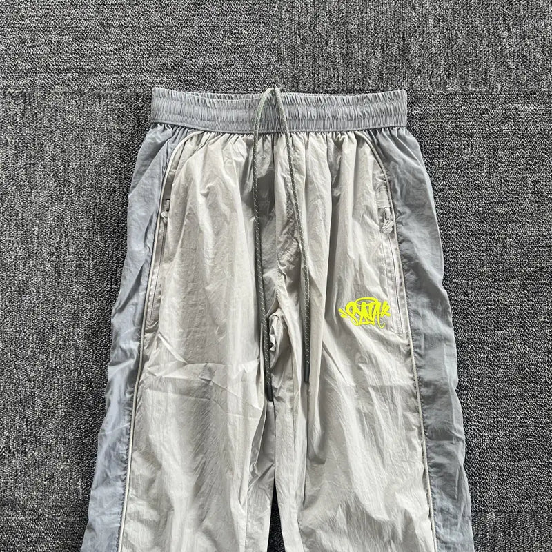 The Syna World Grey Yellow Logo Track Pants feature zip pockets, dark gray side panels, an elastic waistband, and a yellow graffiti-style logo—perfect for elevating your athleisure wardrobe.