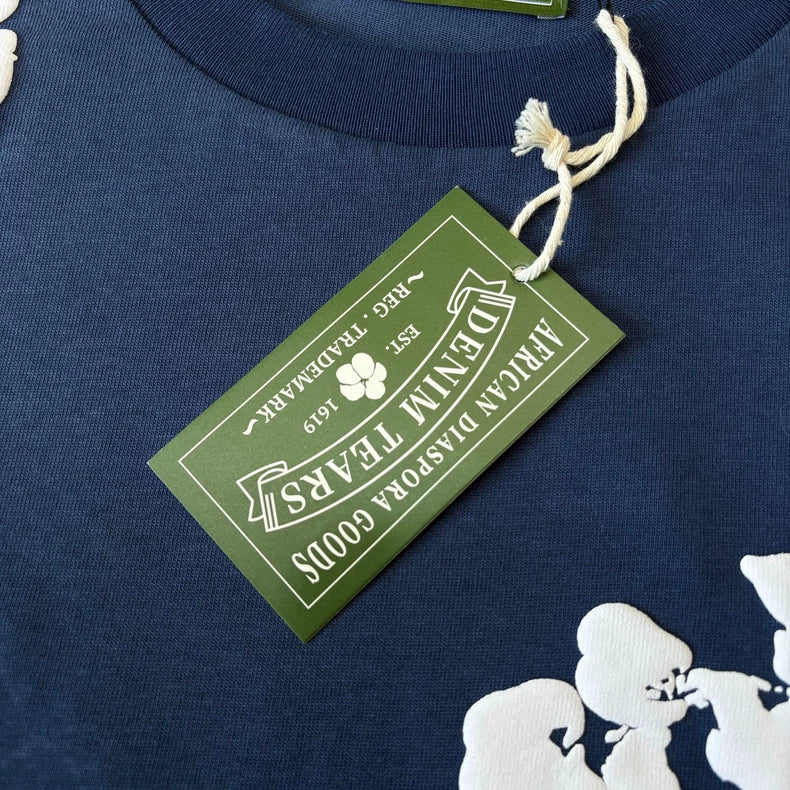 A close-up of a green tag on the Denim Tears Navy Cotton Wreath All-Over Print T-Shirt, reading African Diaspora Goods, Denim Tears, Est. 2019, Reg. Trademark. White floral and cotton wreath graphics appear on the fabric.