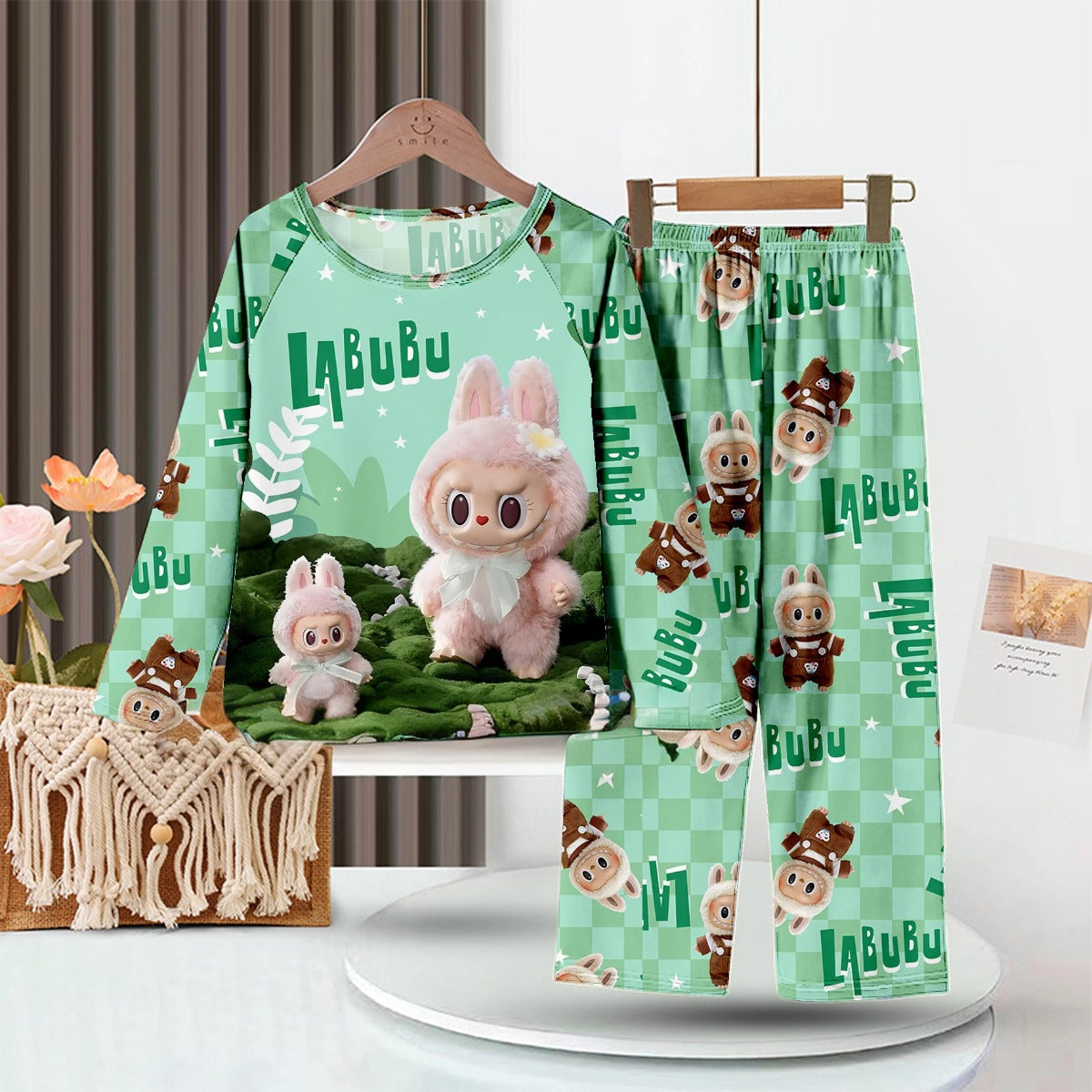 The LABUBU Baby Pajama Set - Cozy Green Milk Silk Sleepwear for Kids, featuring cartoon characters and a bunny theme, includes a long-sleeve shirt and pants. A plush toy and potted plants enhance the woodland aesthetic against a striped wall backdrop.