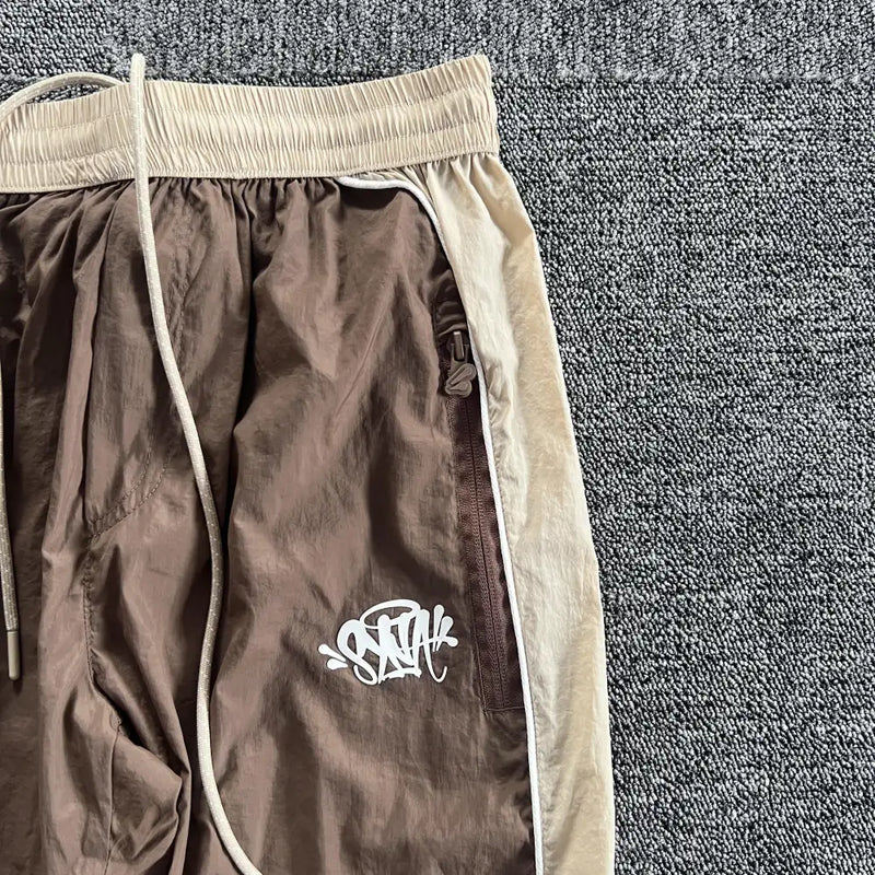 The Syna World Brown Beige Side Stripe Track Pants are soft cotton-polyester joggers with a white graffiti logo on one leg, an elastic waistband, a visible drawstring, and laid on a textured gray carpet.