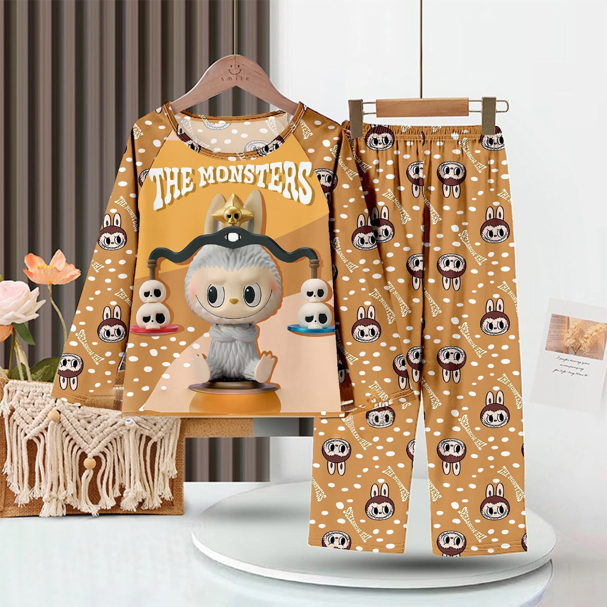 The LABUBU Baby Pajama Set, featuring &quot;The Monsters&quot; theme in brown, is made from soft milk silk with charming character artwork on the top and matching patterned pants. It hangs attractively on a rack near a decorative plant and woven basket.