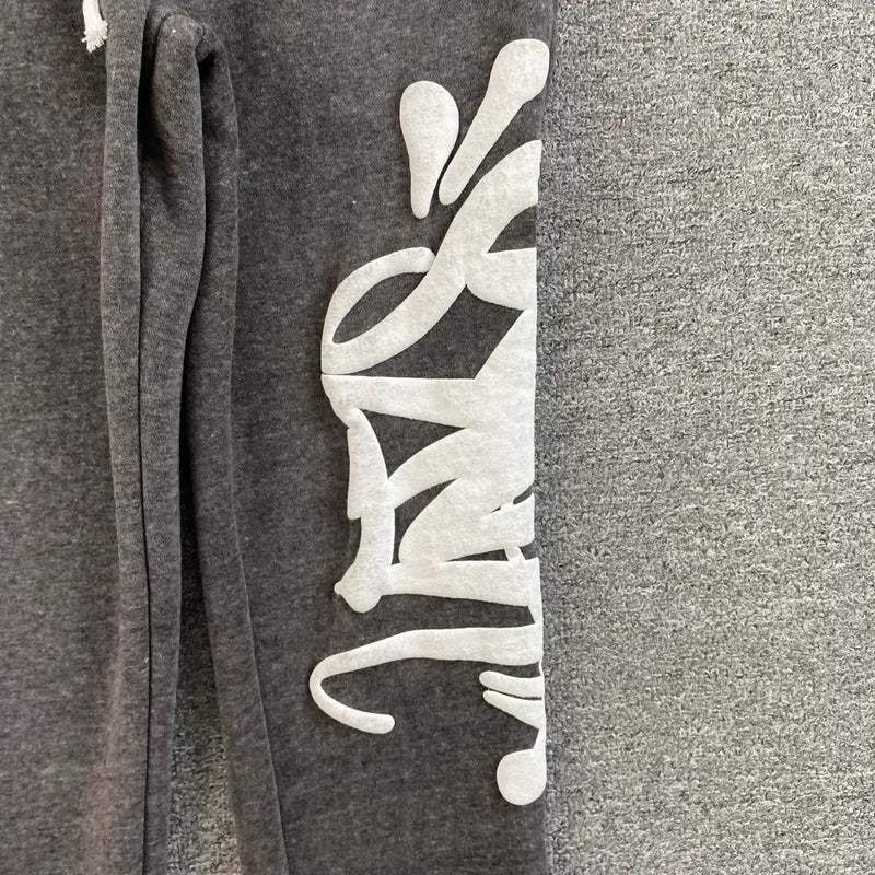 The Syna World Dark Gray Graffiti Letter Print Jogger Pants feature bold white graffiti-style text along one leg, showcased against a textured gray background.