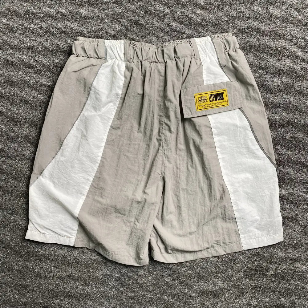 A pair of Corteiz Beige Colorblock Nylon Shorts with Belt, featuring a relaxed fit and colorblock design, laid flat on a gray textured surface.