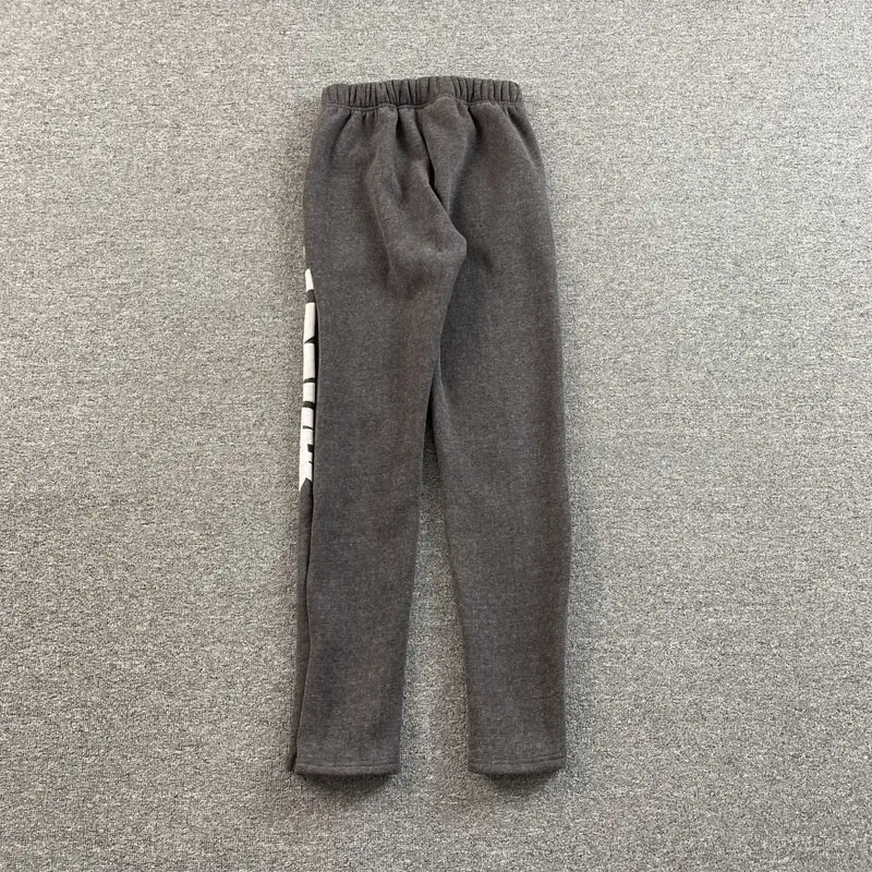 The Syna World Dark Gray Graffiti Letter Print Jogger Pants feature an elastic waistband and bold white lettering on the left leg, offering urban style and comfort, shown laid flat on a textured gray surface.